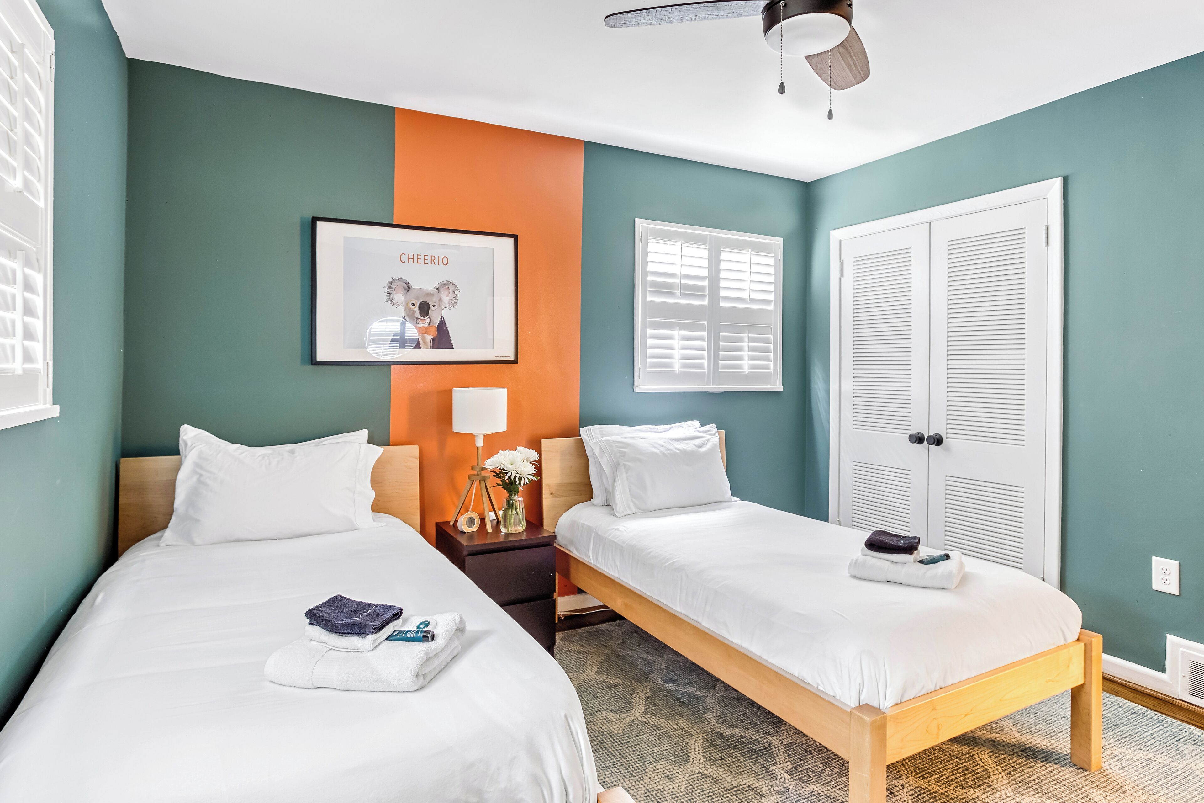 A hotel room with two beds, a framed picture on the wall, a nightstand with a lamp, and a window with shutters.