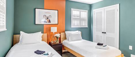 A hotel room with two beds, a framed picture on the wall, a nightstand with a lamp, and a window with shutters.