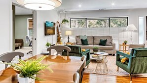 A modern living room with a wooden dining table, green armchairs, and a sofa. There is a TV mounted on the wall, a piano, and a large window with blinds. - Modern and clean home, quick access to attractions (Atlanta)