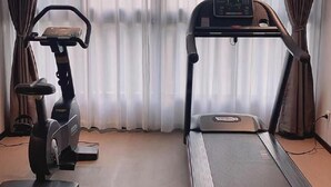 Quarto com esteira e máquina elíptica, janelas amplas com cortinas leves e piso de madeira.