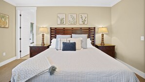 Une chambre avec un grand lit, deux tables de chevet avec des lampes, une porte et trois reproductions encadrées de motifs botaniques accrochées au mur.
