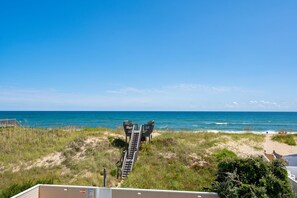 A beach view with a wooden staircase leading to a beach area, a sandy path, and a clear blue sky.