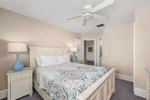 A bedroom with a bed, nightstands, a ceiling fan, and a door leading to another room.
