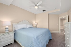 Un dormitorio con cama, mesitas de noche, cómoda, ventilador de techo y una puerta que da a otra habitación.