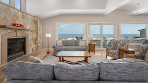 A spacious living room with a fireplace, a sofa set, a coffee table, and a view of the ocean. - Take It to the Limit: 16 Bedrooms, oceanfront, pool, elevator, hot tubs, rec room, theater room! (Nags Head)
