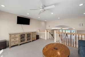 A spacious living room with a ceiling fan, a mounted TV, a wooden coffee table, and a view of the beach through the balcony.
