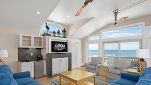 A spacious living room with a large window, a ceiling fan, and a kitchen area with white cabinets and a TV. - One of These Nights: Ocean views, private pool & hot tub, rec room & elevator! (Nags Head)