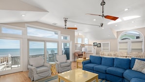 A spacious living room with a large blue sofa, a glass coffee table, and a view of the ocean through large windows. - One of These Nights: Ocean views, private pool & hot tub, rec room & elevator! (Nags Head)