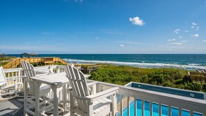 A poolside area with white Adirondack chairs, a wooden deck, and a view of the ocean. - One of These Nights: Ocean views, private pool & hot tub, rec room & elevator! (Nags Head)