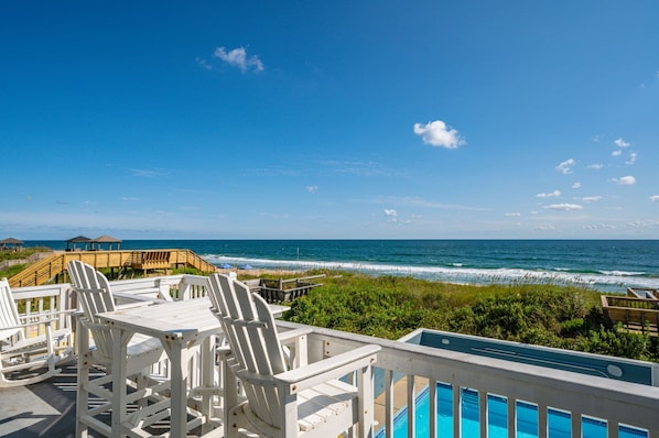 A poolside area with white Adirondack chairs, a wooden deck, and a view of the ocean.