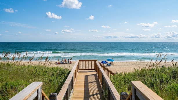 A wooden boardwalk leading to a beach with people and umbrellas. - One of These Nights: Ocean views, private pool & hot tub, rec room & elevator! (Nags Head)