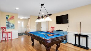 Une table de billard dans une pièce dont le sol est recouvert de parquet, avec un téléviseur fixé au mur et un tableau accroché au mur.