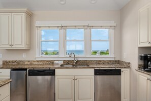 A kitchen with white cabinets, stainless steel appliances, a granite countertop, and a view of the ocean through a window. - Tequila Sunrise-Oceanfront w/views, private pool, hot tub, rec room w/pool table (Nags Head)
