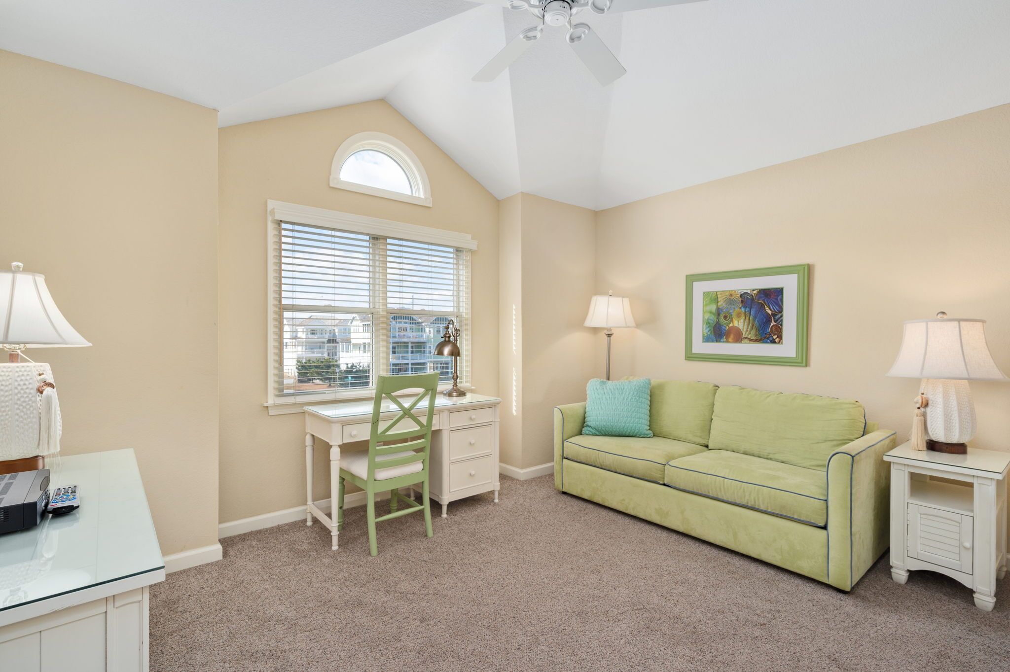A room with a desk, a green couch, a window with blinds, a ceiling fan, and a framed picture on the wall.