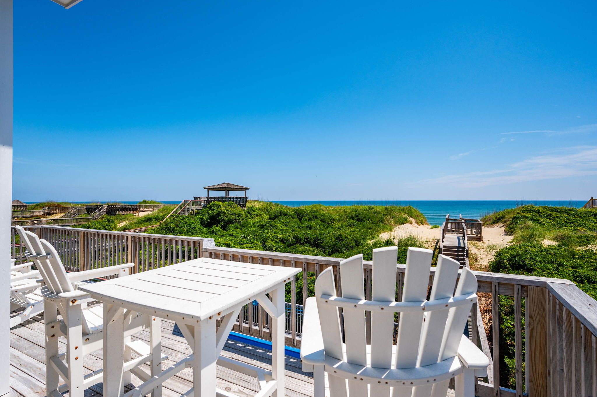 A wooden deck with white chairs and a table, overlooking a beach with a wooden walkway and a gazebo.