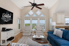 A spacious living room with a large window, a ceiling fan, and a flat-screen TV mounted on the wall. - Tequila Sunrise-Oceanfront w/views, private pool, hot tub, rec room w/pool table (Nags Head)