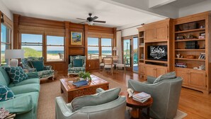 A spacious living room with large windows, a ceiling fan, and wooden furniture. - Gillam Hall: Oceanfront, private walkway, tons of space and beautiful beaches. (Nags Head)
