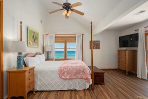 A bedroom with a bed, a ceiling fan, a TV, a wooden dresser, and a view of the ocean.