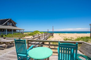 A wooden deck with a table and chairs, overlooking a beach with a clear sky.