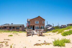 A beachfront house with a sandy beach in front, a wooden deck, and a flag.