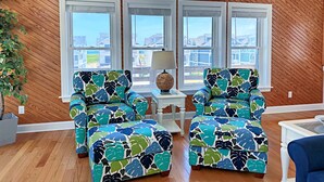 A room with wooden walls, a large arched window, and two matching armchairs with a tropical leaf pattern. - Phelps Brothers Too:Ocean Views, Private pool, hot tub, elevator & Dog Friendly! (Nags Head)
