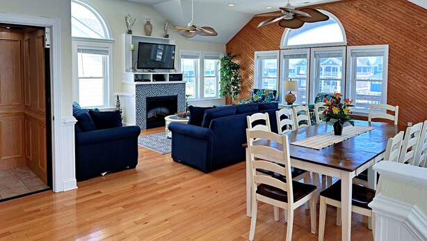 A spacious living room with a fireplace, a dining area with a wooden table and chairs, and a large window with a view of houses. - Phelps Brothers Too:Ocean Views, Private pool, hot tub, elevator & Dog Friendly! (Nags Head)