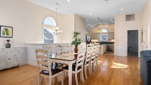 A spacious dining area with a long wooden table and white chairs, a potted plant, and a large window providing natural light.