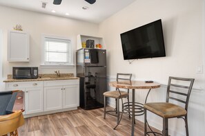 Cocina compacta con armarios blancos, microondas, refrigerador, ventana con persianas, televisor montado en la pared y un pequeño comedor con dos sillas y una mesa.