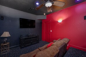 Una habitación con pared roja, ventilador de techo, televisor de pantalla plana, mueble de madera oscura, alfombra estampada y sofá.
