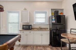 A kitchen with white cabinets, a microwave, a refrigerator, a pool table, and a dining table with chairs.