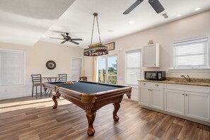 A pool table in a room with wooden flooring, white cabinets, and a ceiling fan.