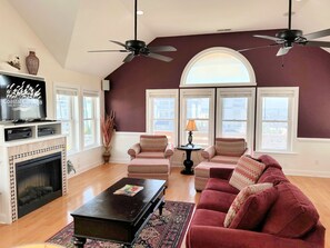 A living room with a fireplace, two ceiling fans, a television, and a large window with a view.