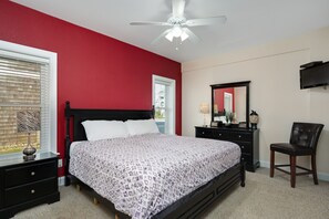 Quarto com parede vermelha, estrutura de cama preta, ventilador de teto, cômoda, cadeira e TV fixada na parede.
