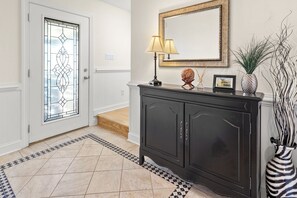 A hallway with a patterned floor, a dark wooden console table with a lamp, a framed mirror, and a vase with plants.