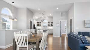 A spacious kitchen with white cabinetry, a dining area with a wooden table and white chairs, and a blue sofa in a living area. - Phelps: Semi-oceanfront w/private pool, hot tub, rec and media room & elevator! (Nags Head)