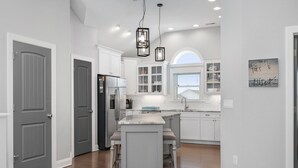 A modern kitchen with white cabinetry, a central island with bar stools, pendant lighting, and a view to the outside. - Phelps: Semi-oceanfront w/private pool, hot tub, rec and media room & elevator! (Nags Head)