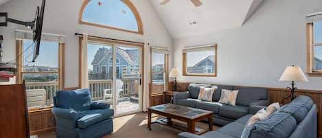 A spacious living room with a high ceiling, a ceiling fan, a large window with a view, and a sliding glass door leading to an outdoor area.