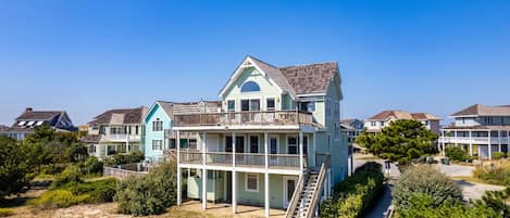 A multi-story beachfront house with a balcony, a staircase leading to the upper floor, and a wooden deck.
