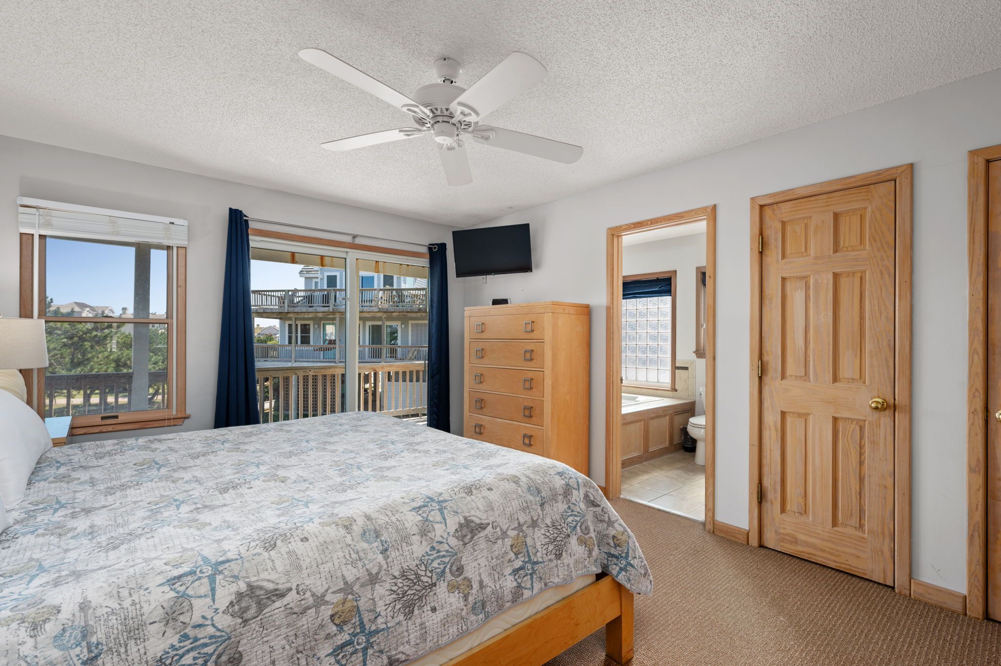 Une chambre à coucher avec un lit, un ventilateur de plafond, une télévision, une armoire et une salle de bain visible à travers une porte ouverte.