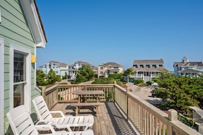 A wooden deck with white chairs and a picnic table, overlooking a row of houses with a clear blue sky above.