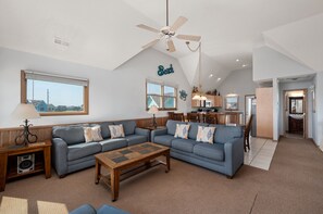 A spacious living room with a large sofa set, a coffee table, and a ceiling fan. A kitchen area is visible in the background.