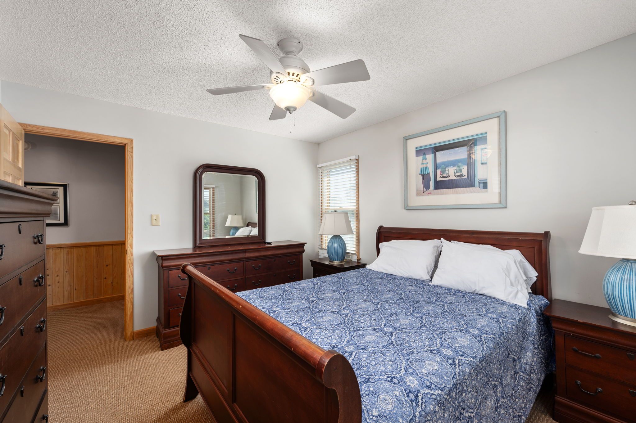 A bedroom with a wooden bed, a ceiling fan, a dresser, a nightstand, a lamp, a framed picture on the wall, and a door leading to another room.