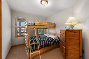 A bunk bed with a striped bedspread, a wooden dresser, and a window with blinds.