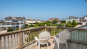 Una terraza de madera con dos sillas blancas, con vista a una zona residencial costera con casas y vegetación.