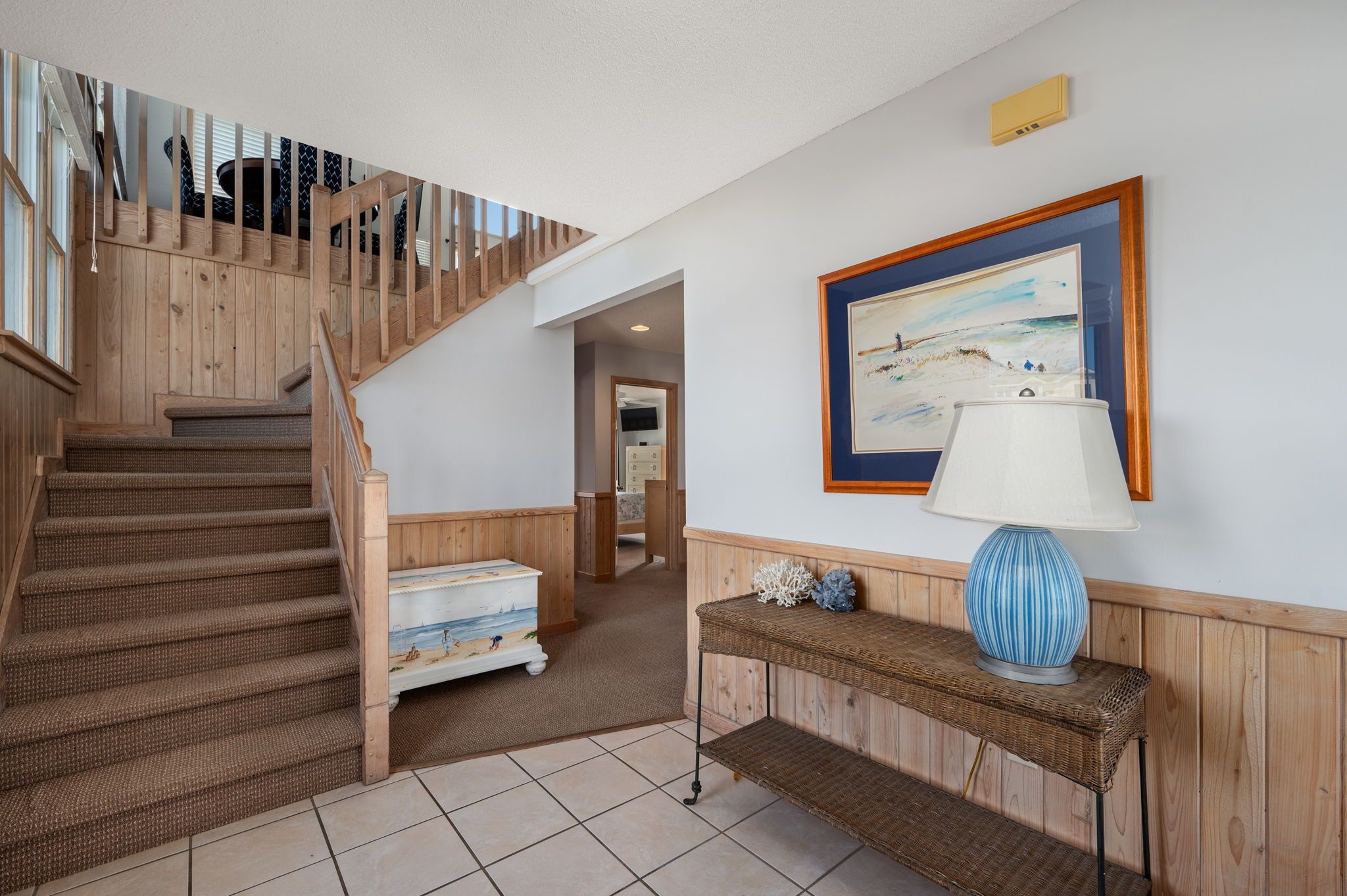 Un escalier avec des rampes en bois menant à un palier, un tableau encadré accroché au mur et une console en osier sur laquelle est posée une lampe.