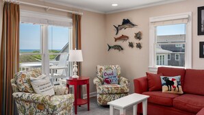 A living room with a view of the ocean, a red sofa, a patterned armchair, and a white table. - At Ease: Ocean views, short walk to the beach, close to fishing pier. (Nags Head)