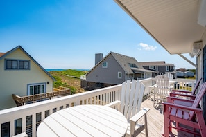 A deck with white chairs and a table overlooking a beach and houses.
