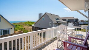 A wooden deck with white railings, a table, and chairs overlooking a beach and houses. - At Ease: Ocean views, short walk to the beach, close to fishing pier. (Nags Head)