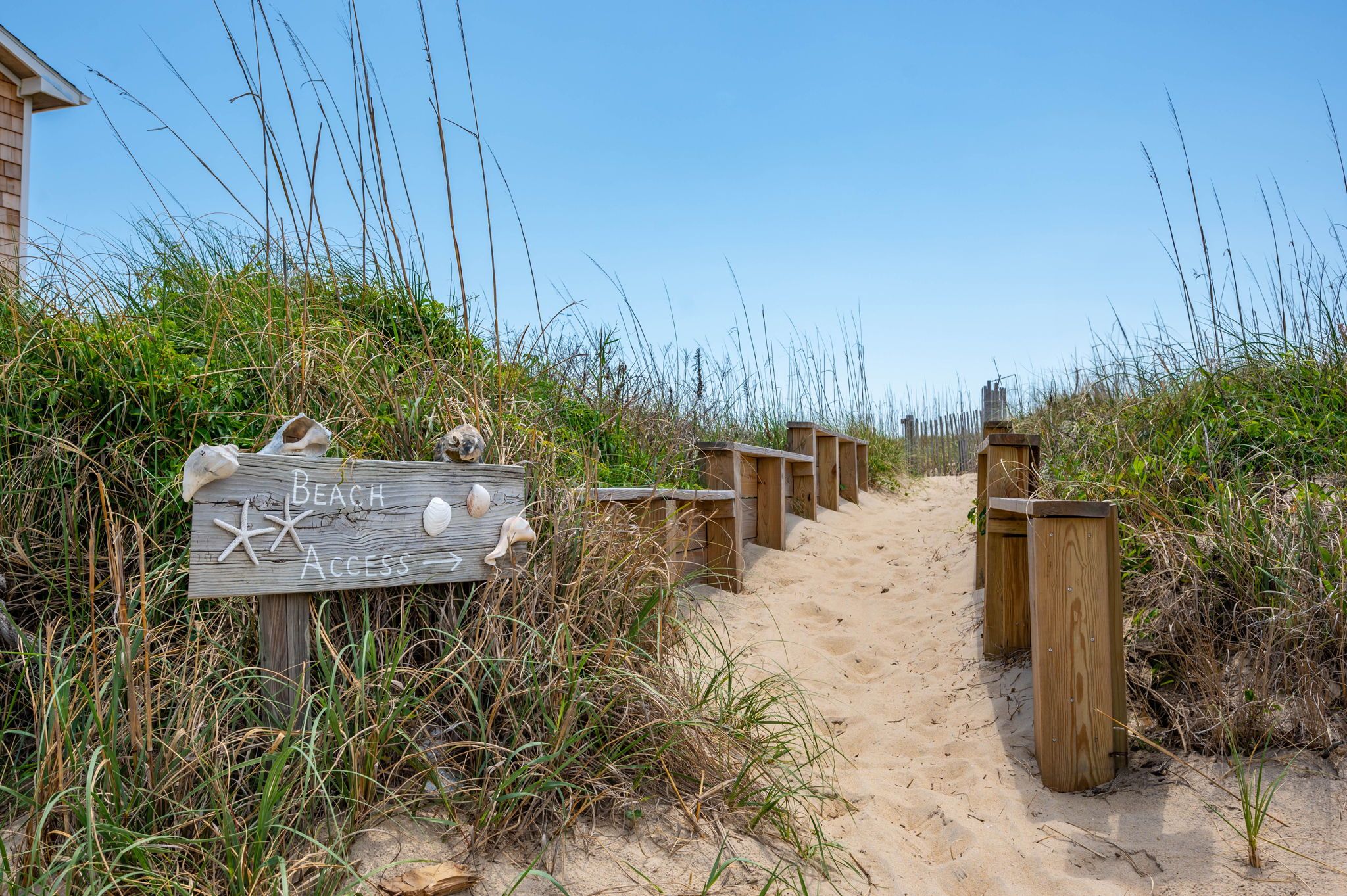 A wooden sign with 'Beach Access' and starfish decorations, leading to a sandy path with wooden barriers.