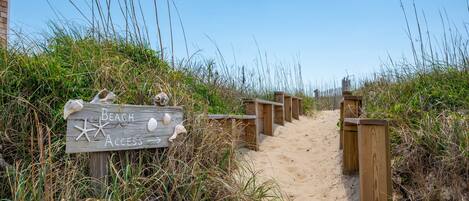 A wooden sign with 'Beach Access' and starfish decorations, leading to a sandy path with wooden barriers.